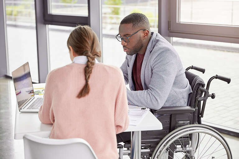 A black man using a wheelchair collaborates at a desk and computer with a young, brown-haired woman. They look like they are working on a plan together.