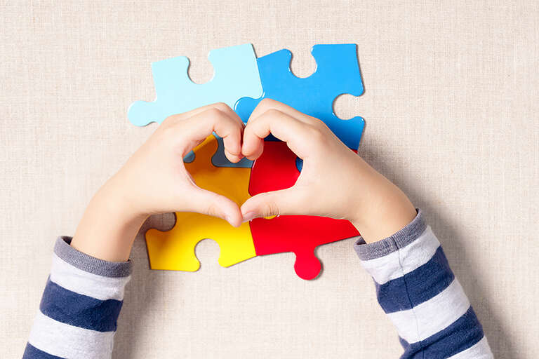 A child's hands making a heart over pieces of a puzzle that often represent the symbol and colors for Autism.